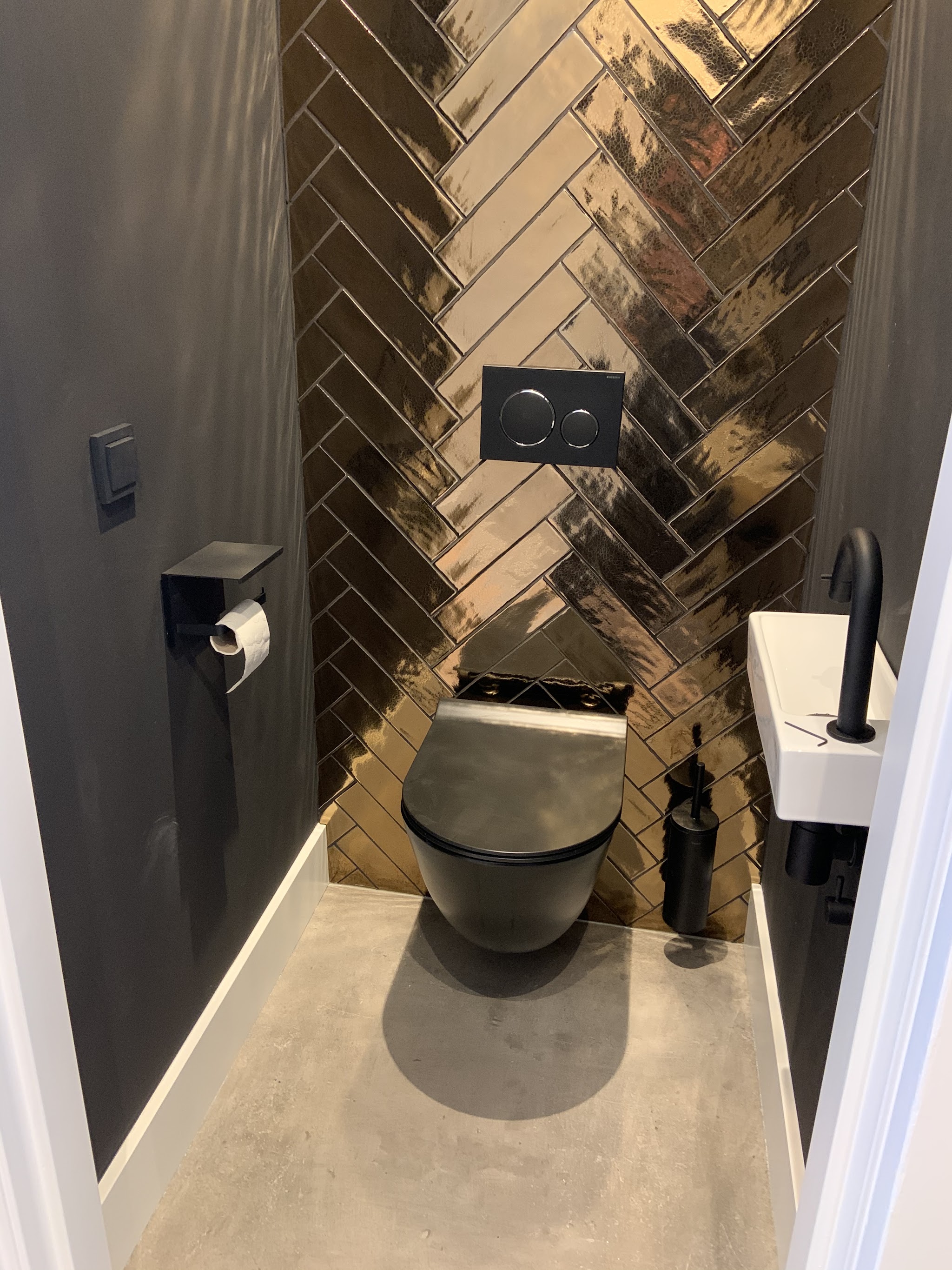 Toiletruimte met zwart hangtoilet, wand met goudkleurige visgraattegels en een klein fonteintje